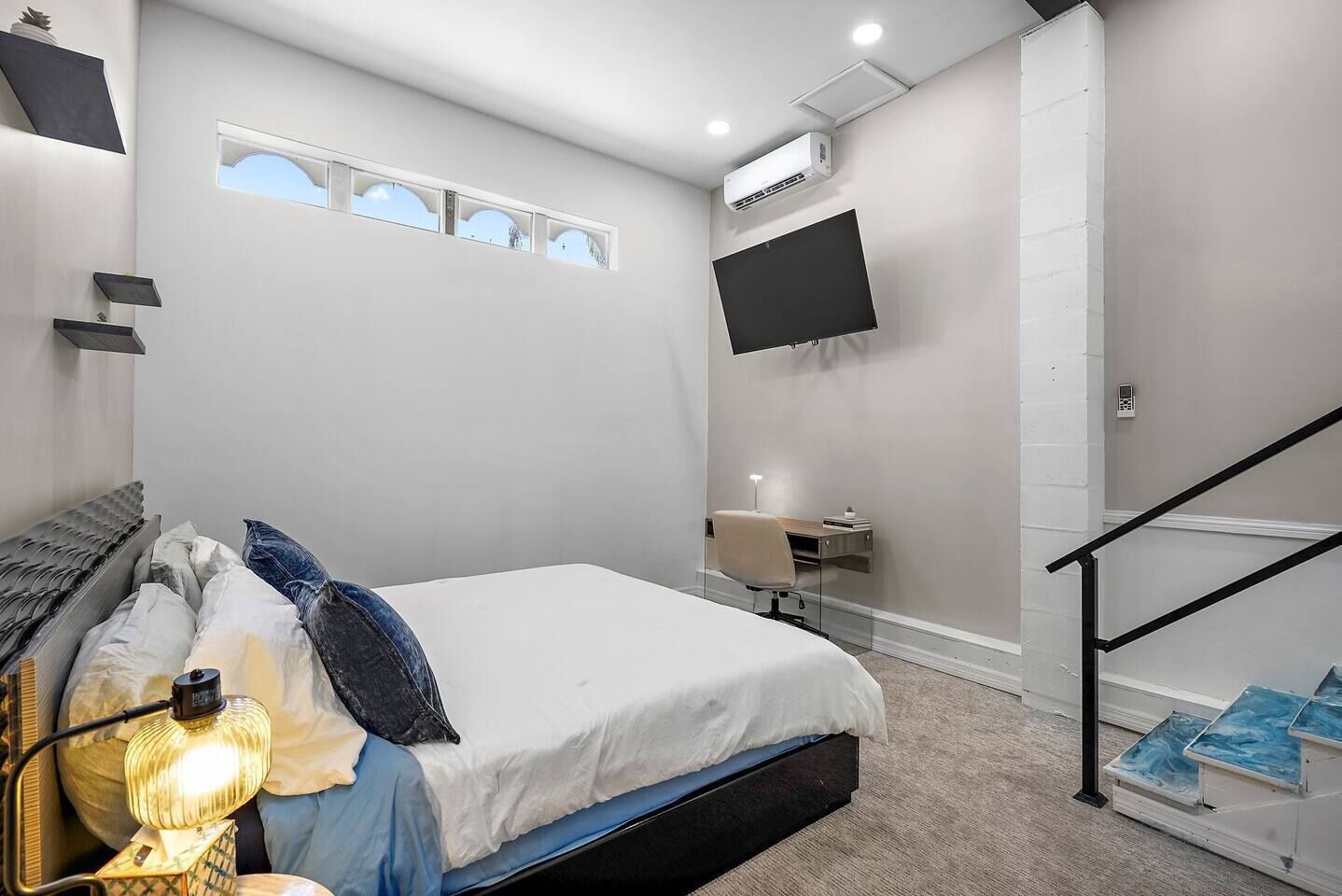 A bedroom with a bed, bedside lamp, and a desk with a chair. There is a TV mounted on the wall and a window with a view.
