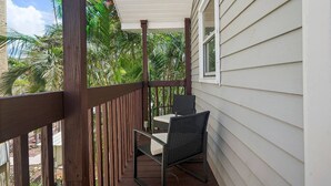 A covered balcony with a table and chairs, surrounded by a wooden railing, overlooking a lush garden with palm trees. - Beach Bliss Veranda Unit 1 (Redington Shores)