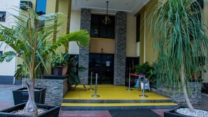 Entrada de hotel con alfombra amarilla, plantas en macetas y una puerta negra.