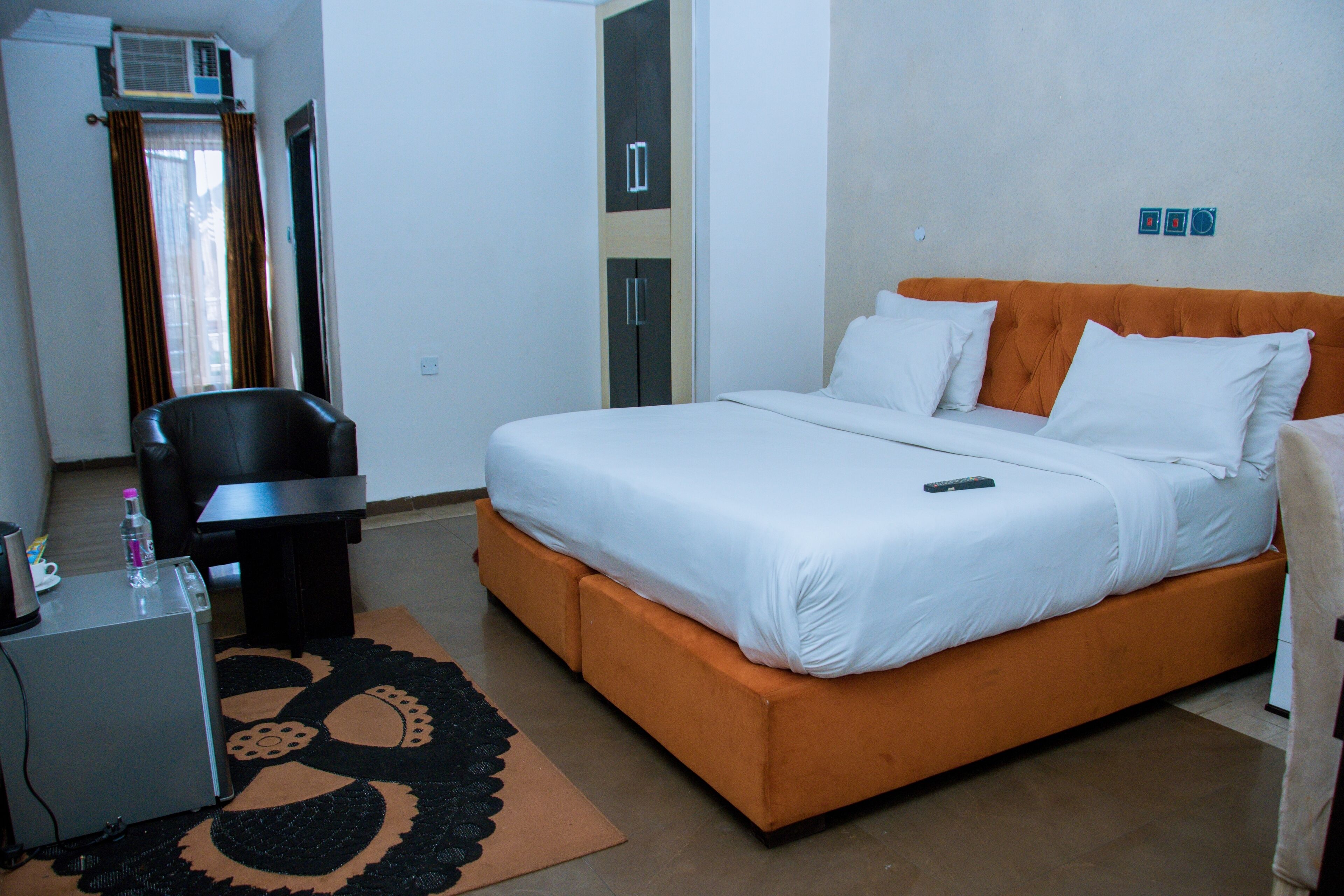 A hotel room with a bed, a desk, a chair, and a small table with a bottle.