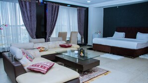 Una habitación de hotel moderna con sofá, mesa de centro de vidrio, cama y un ventanal con cortinas.
