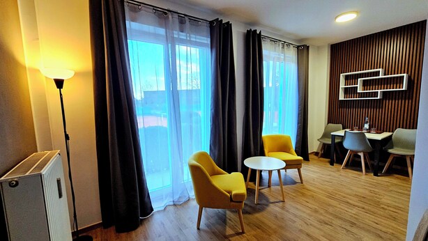Une chambre d’hôtel moderne avec un plancher en bois, deux fauteuils jaunes, une petite table ronde, un radiateur blanc et une grande fenêtre avec des rideaux noirs.