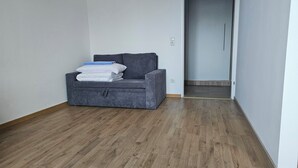 Une petite chaise de couleur gris foncé, recouverte de tissu, avec un coussin et des couvertures pliées, placée contre un mur blanc dans un couloir étroit au plancher en bois.