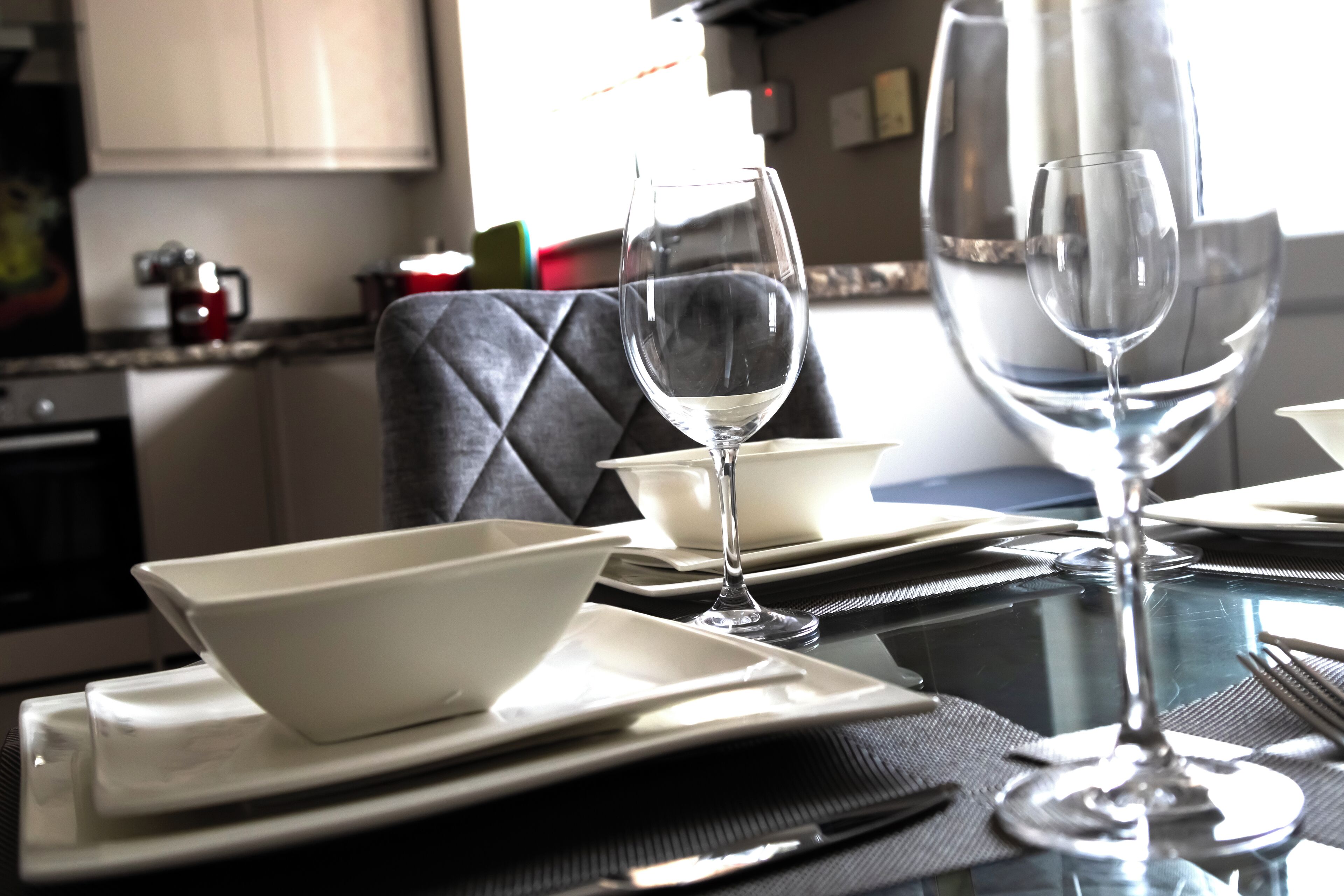 A dining table set with white plates, bowls, and wine glasses, with a kitchen in the background.