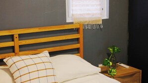 Ein ordentlich bezogenes Bett mit Holz-Kopfteil, ein Nachttisch mit Pflanze und ein Fenster mit Jalousien.