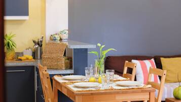 A dining area with a wooden table set for two, a kitchen in the background, and a brown sofa with a yellow pillow.