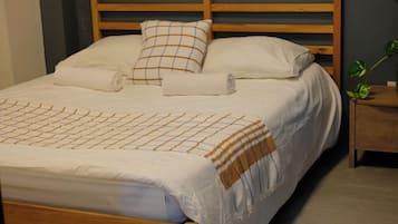 A single bed with a wooden frame, a bedside table with a plant, and a window with blinds.