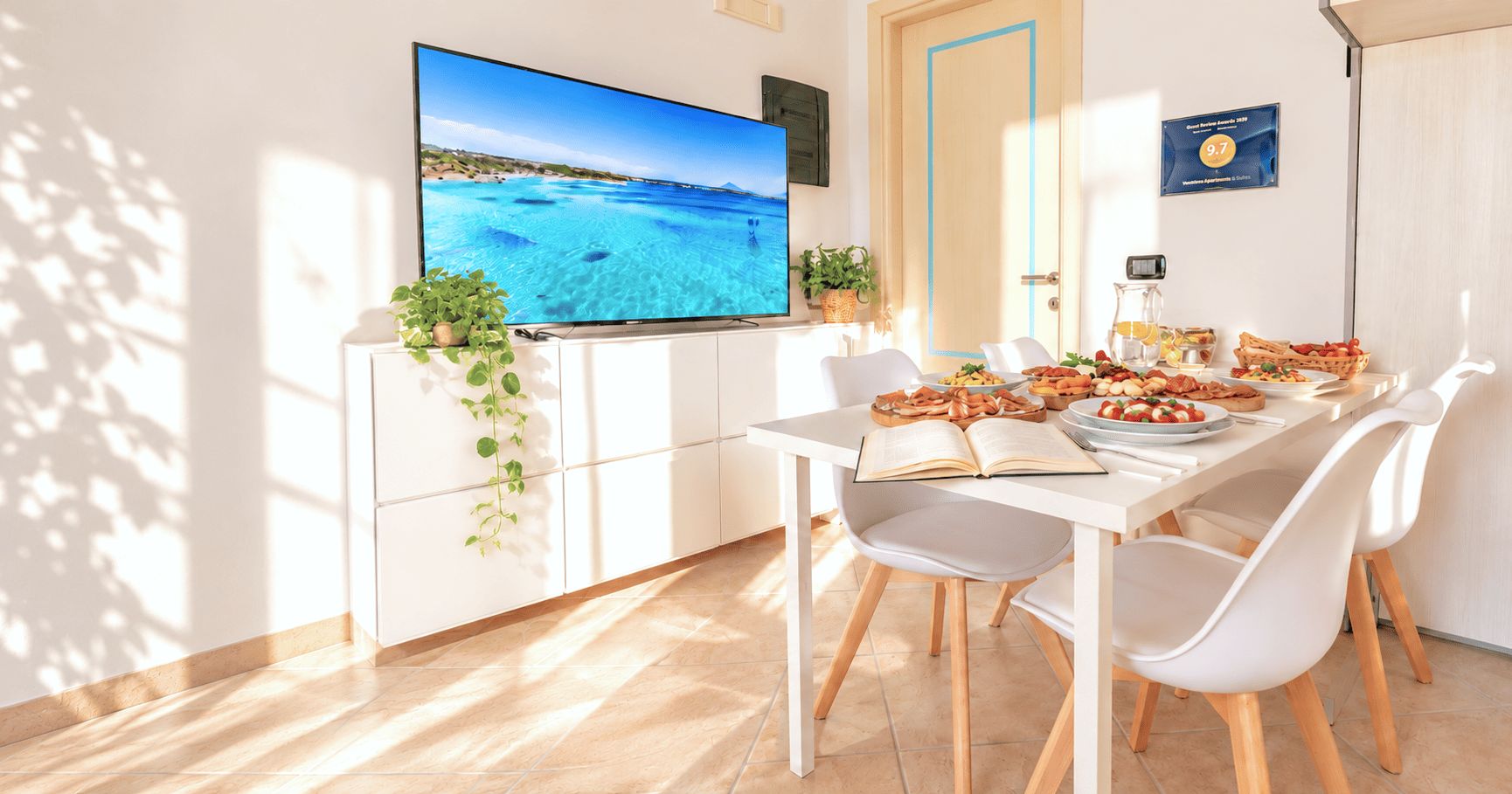 A modern dining area with a flat-screen TV, a white table set for two, and a view of a beach through the window.