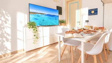 Un comedor moderno con un televisor de pantalla plana, una mesa blanca dispuesta para dos personas y vistas a una playa a través del ventanal.