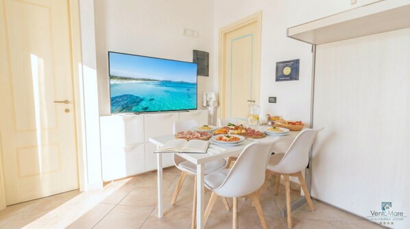 A dining area with a table set for four, a television mounted on the wall, and a view of the sea. - Vento Mare Apartments & Suites-Palau (Palau)