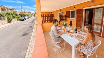 Un comedor con una mesa dispuesta para dos personas, vista a la calle y un edificio con paredes naranjas y contraventanas de madera.