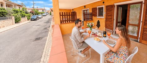Un coin repas avec une table dressée pour deux personnes, une vue sur la rue et un bâtiment aux murs orange et aux volets en bois.