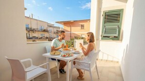 Een balkon met een tafel en stoelen, een raam met luiken en uitzicht op gebouwen.