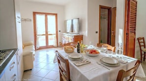 A dining area with a table set for four, a television in the background, and a kitchen area with a stove and cabinets. - Vento Mare Apartments & Suites-Palau (Palau)