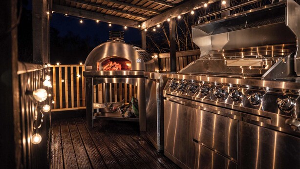 An outdoor kitchen with a built-in grill and oven, surrounded by a wooden deck and string lights. - Family Bungalow, Mountain Views, Pets Welcome (Pontsticill)