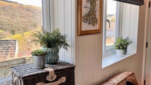 A cozy room with a view of a hilly landscape, a wicker basket with plants, a framed map, and a brown leather sofa. - Family Bungalow, Mountain Views, Pets Welcome (Pontsticill)