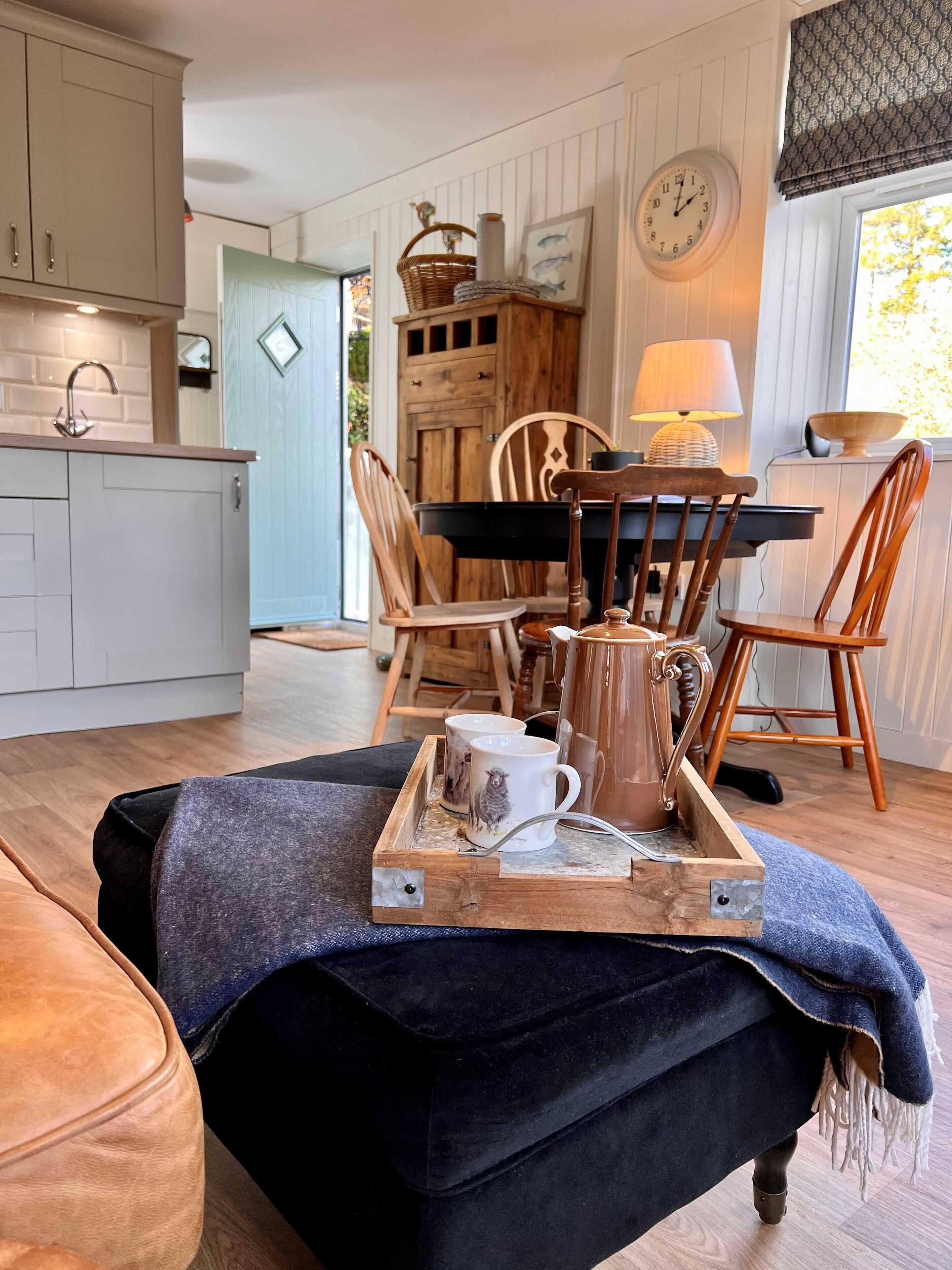 A cozy kitchen with wooden cabinets, a dining table with chairs, a coffee table with mugs and a coffee pot, and a clock on the wall.