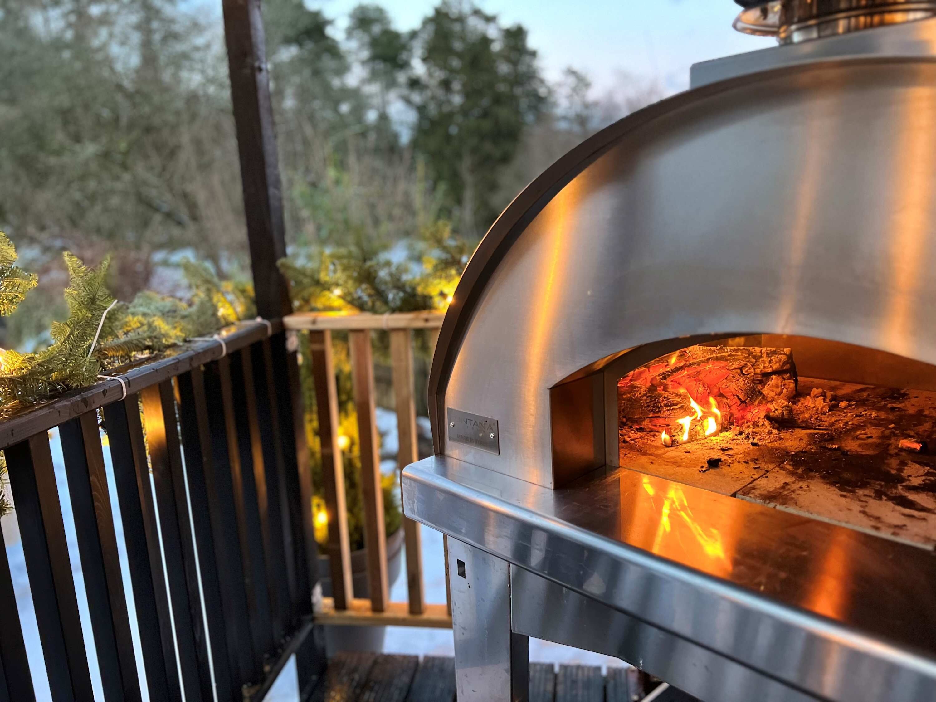 A pizza oven with a fire inside, located outdoors with a wooden deck and greenery in the background.