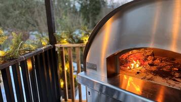 A pizza oven with a fire inside, located outdoors with a wooden deck and greenery in the background.