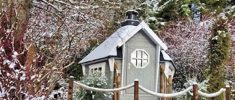 A small, rustic cabin with a steeply pitched roof and a chimney, surrounded by snow-covered trees and a rope barrier.