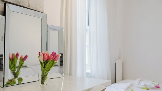 A hotel room with a white bed, a bedside table with a vase of pink and white tulips, and a large mirror on the wall. - 3BR Tower Bridge Parking WiFi (London)