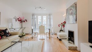 A modern living room with a sofa, coffee table, and a fireplace. There is a dining area with a table and chairs, and a large window with curtains. - 3BR Tower Bridge Parking WiFi (London)