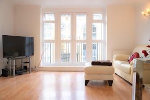 A living room with a flat-screen TV on a stand, a sofa, and a window with a view of buildings. - Gated SE1 Mews House | Free Parking & Balcony (London)