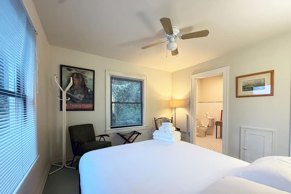 A hotel room with a large bed, a ceiling fan, a chair, a desk, a window with blinds, and a bathroom visible through an open door.