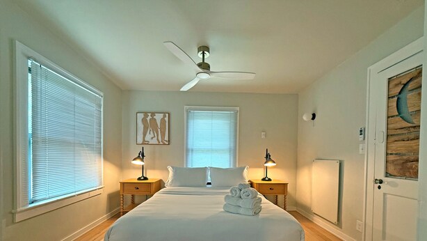 A bedroom with a bed, two bedside tables, a ceiling fan, and two windows with blinds. - Moby Dick Hotel (Ocean Park)