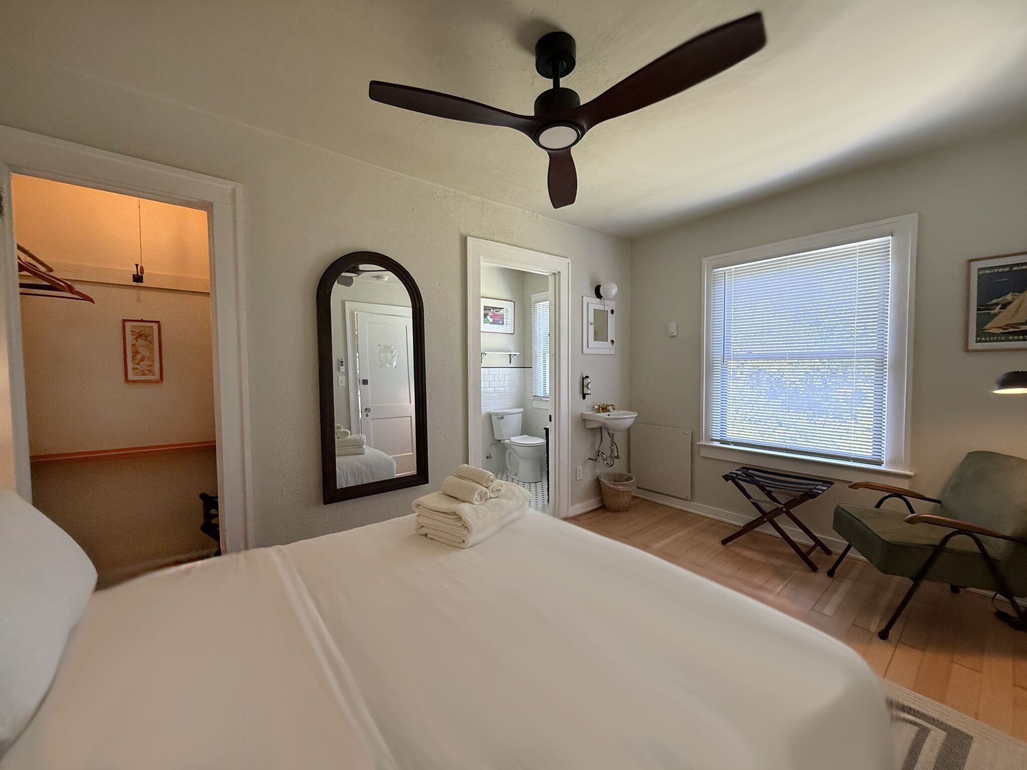A bedroom with a bed, a ceiling fan, a window with blinds, a mirror, and a bathroom visible through an open door.