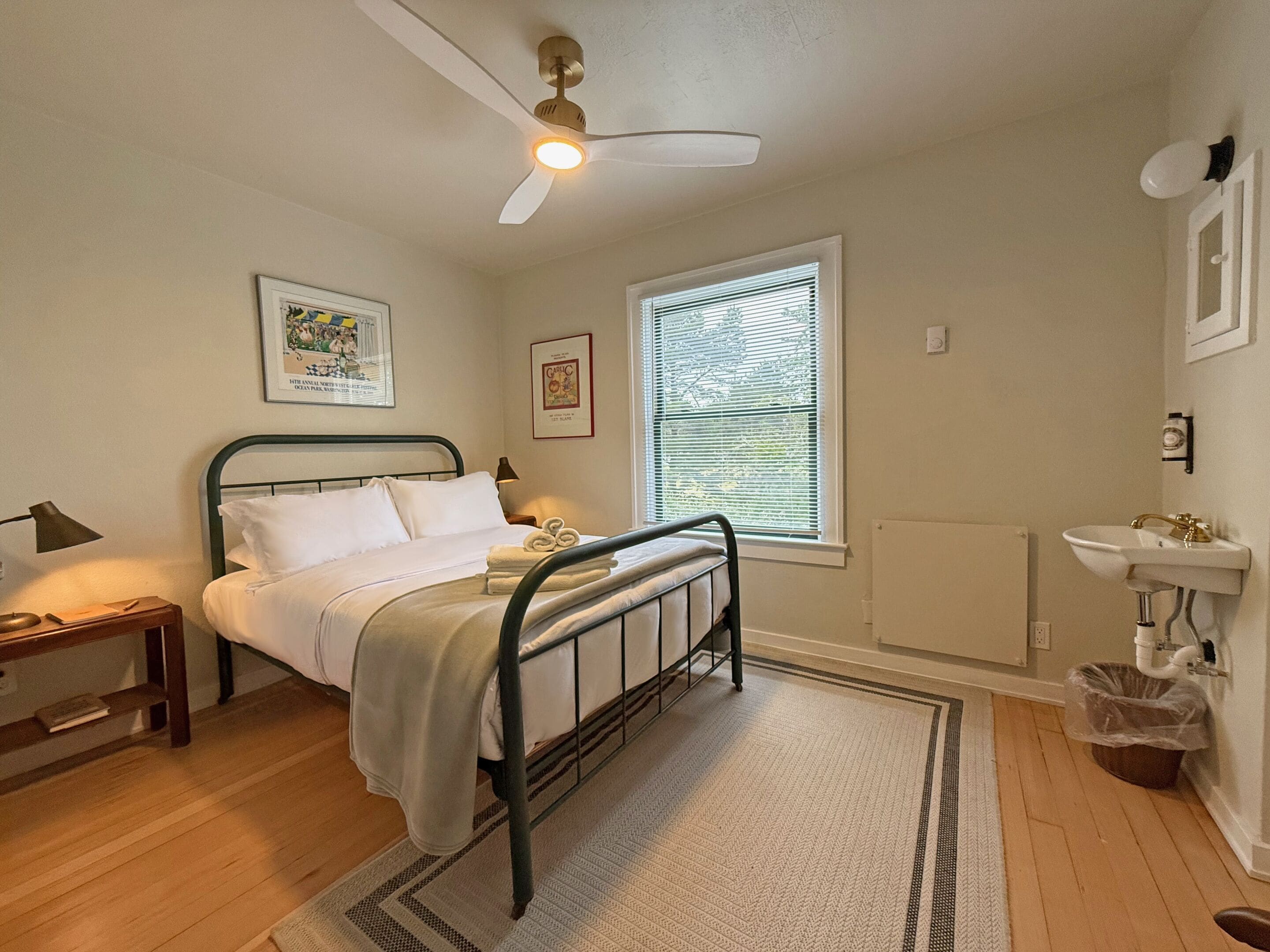 A bedroom with a bed, bedside table, lamp, wall art, window with blinds, ceiling fan, and a bathroom with sink and toilet.