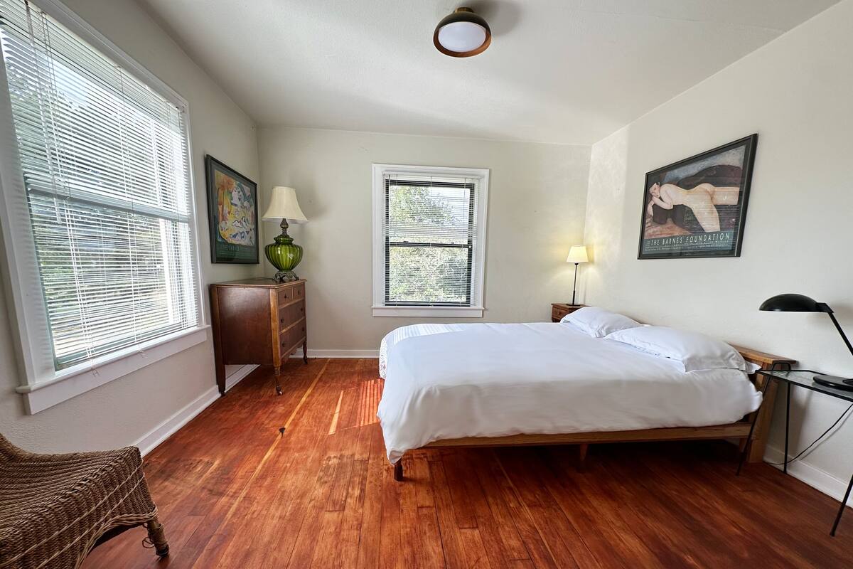 A bedroom with a wooden floor, a bed, a nightstand, a lamp, a chair, and a window with blinds.