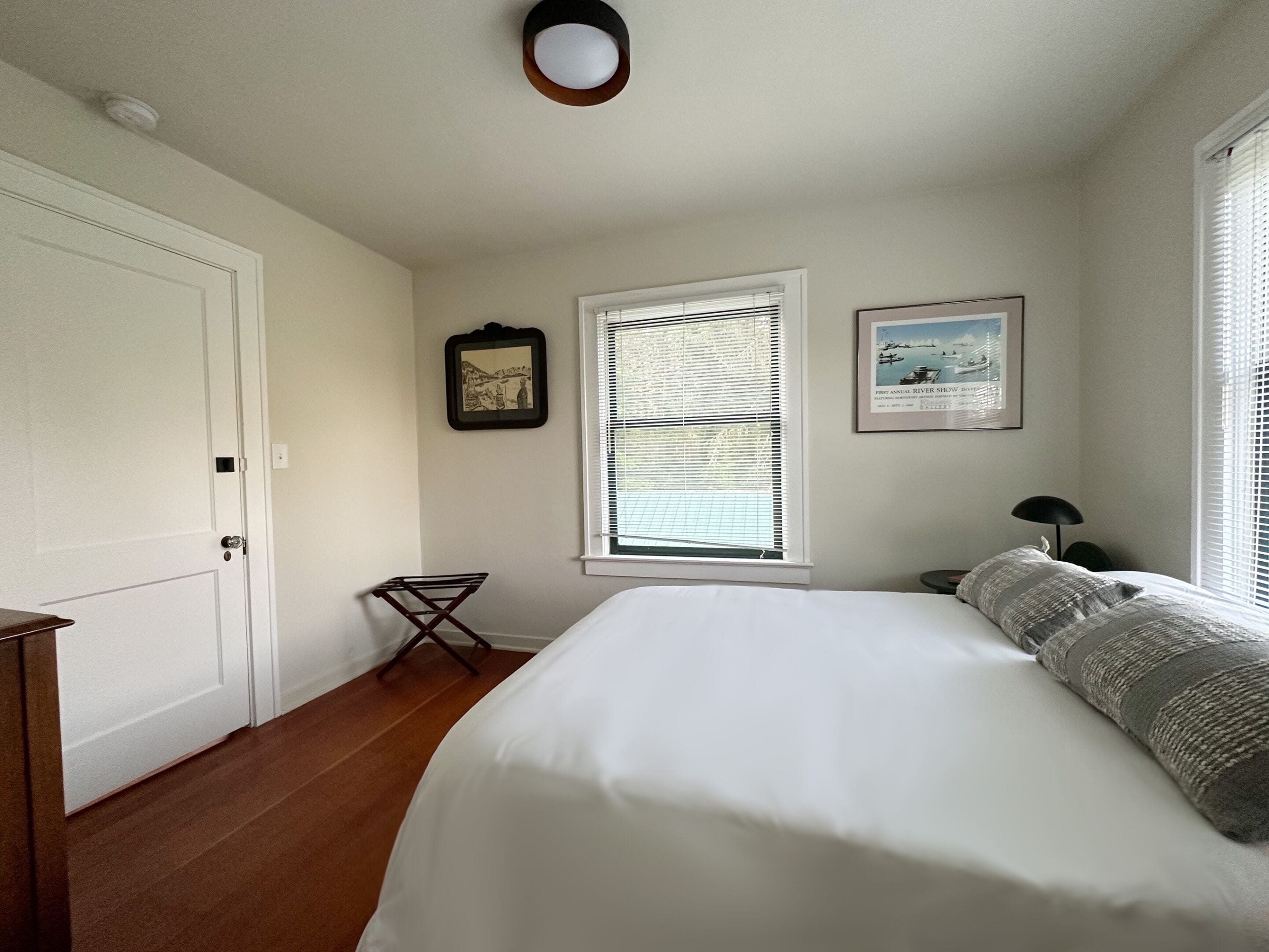 A bedroom with a bed, a window with blinds, a door, a framed picture, and a small table with a lamp.