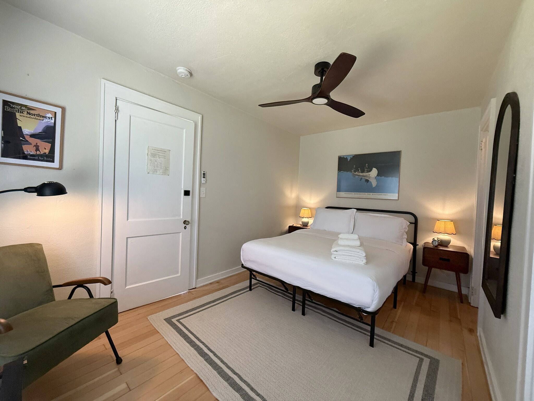 A hotel room with a bed, a ceiling fan, a nightstand with a lamp, a chair, a framed picture on the wall, and a door.