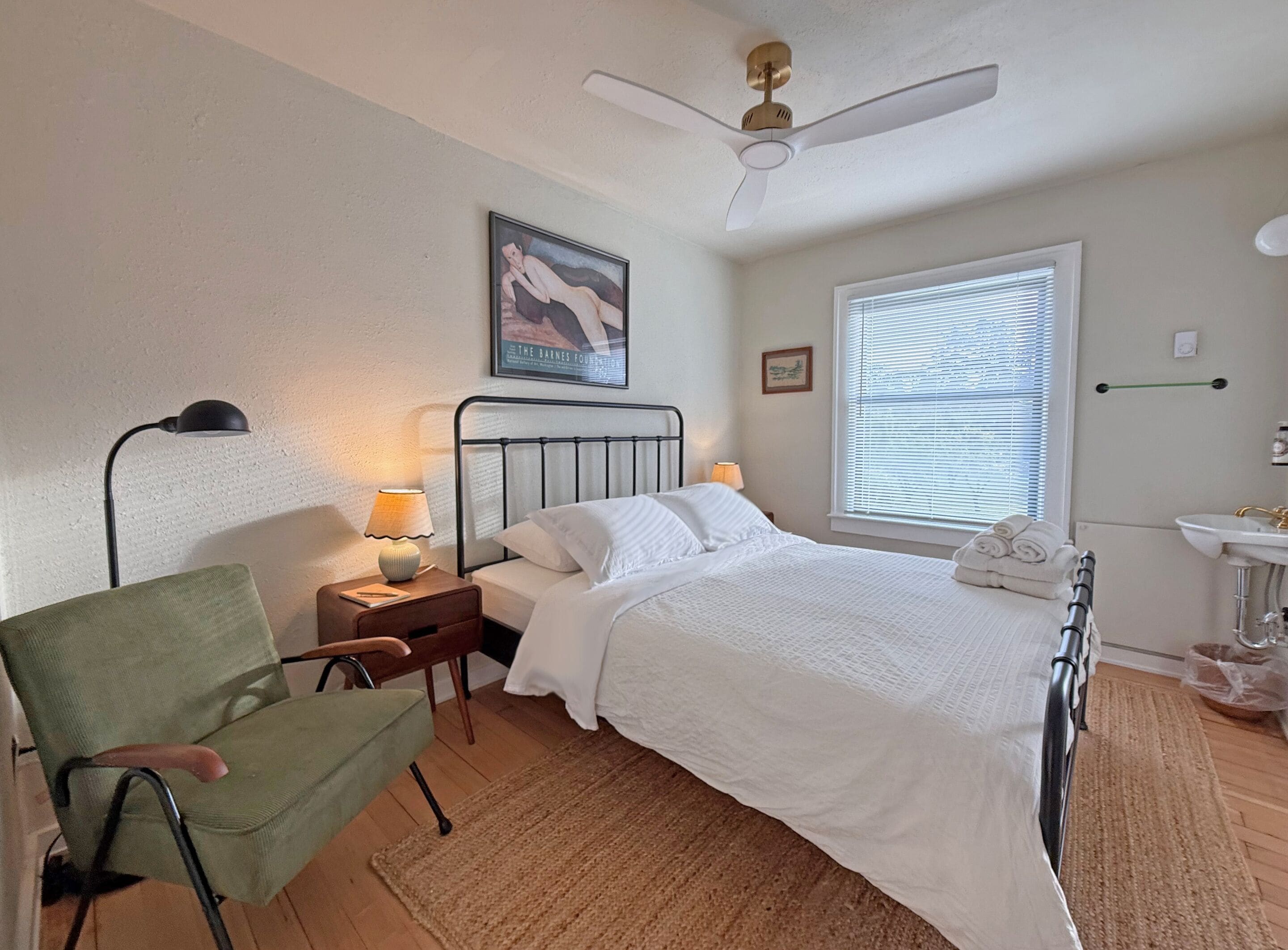 A bedroom with a bed, a chair, a bedside table with a lamp, a ceiling fan, a window with blinds, and a painting on the wall.