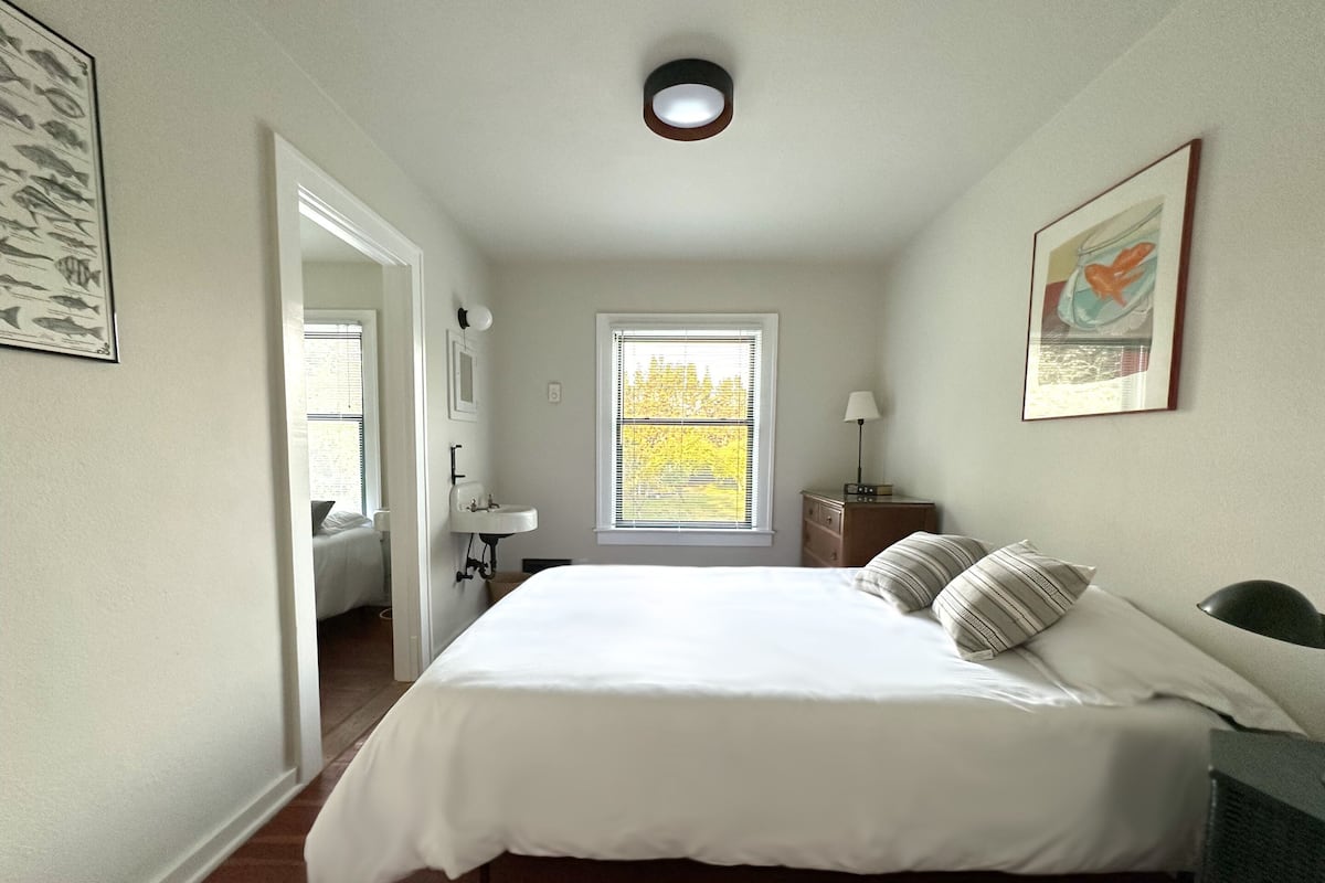 A bedroom with a bed, a nightstand, a window with blinds, a door leading to another room, and a wall-mounted light fixture.