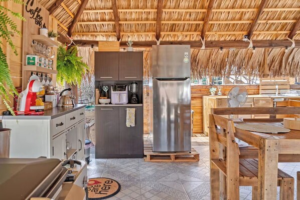 A rustic kitchen with a thatched roof, wooden cabinets, a stainless steel refrigerator, and a coffee bar.