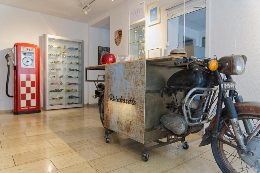 Une moto ancienne, avec un coffre en métal rouillé portant l’inscription « Reinhardts », se trouve dans une pièce où l’on aperçoit une pompe à essence rouge et une vitrine en verre contenant des modèles de voitures.