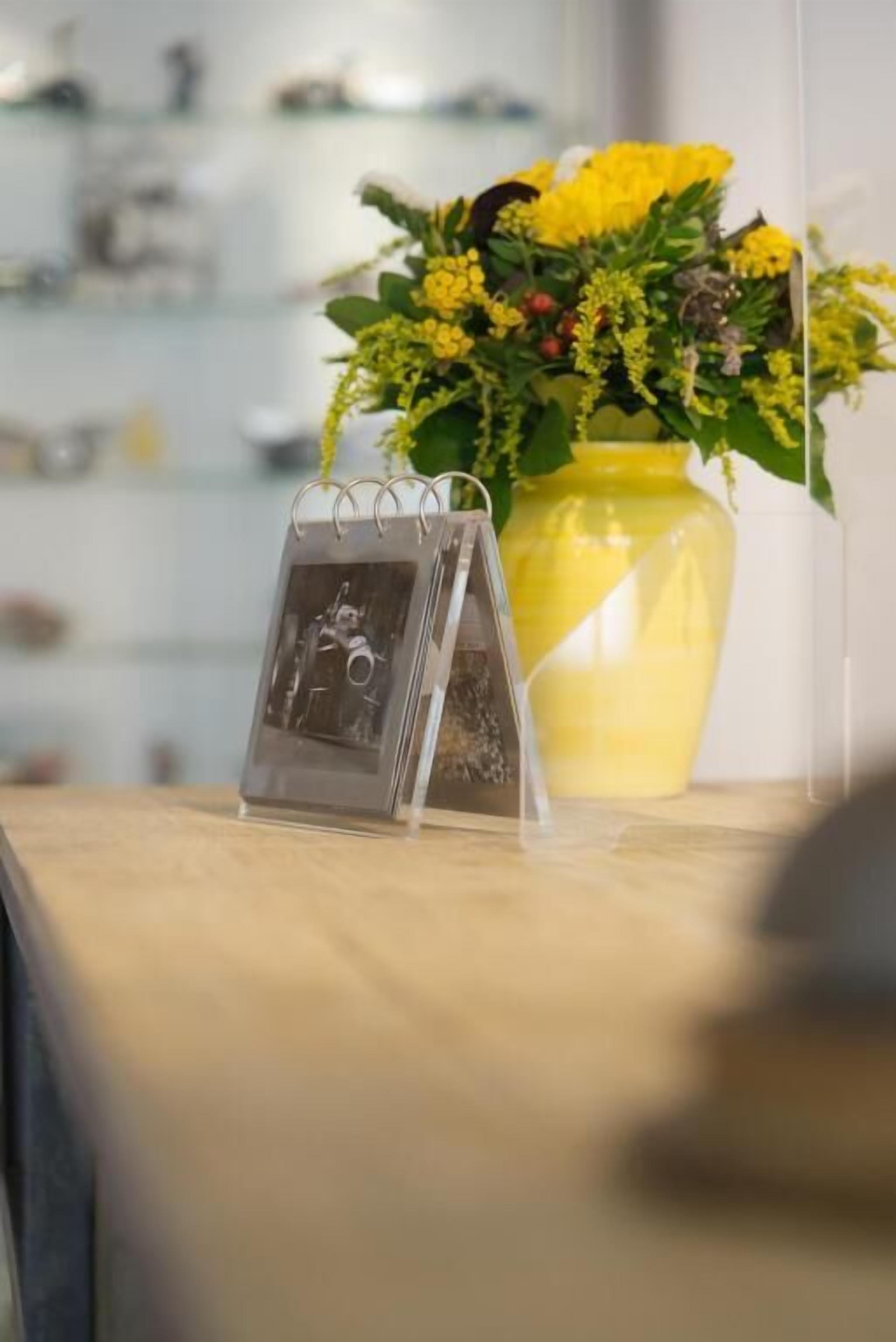 A clear acrylic stand with a photo, a yellow vase with yellow flowers, and a blurred background.
