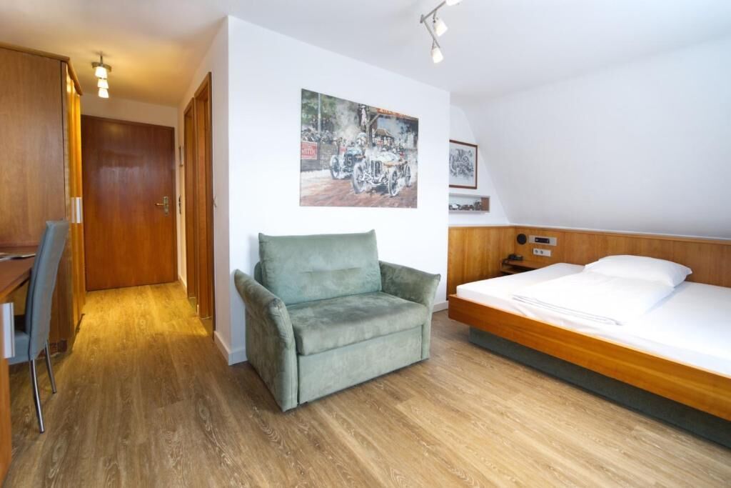 Ein Hotelzimmer mit Holzboden, einem grünen Sessel, einem Bett mit weißer Bettwäsche und einem Gemälde an der Wand.