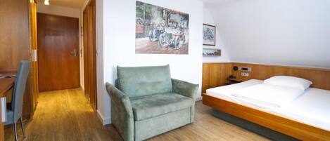 A hotel room with a wooden floor, a green armchair, a bed with white linens, and a painting on the wall.