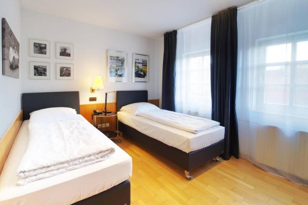 A hotel room with two beds, a nightstand with a lamp, and a large window with curtains.