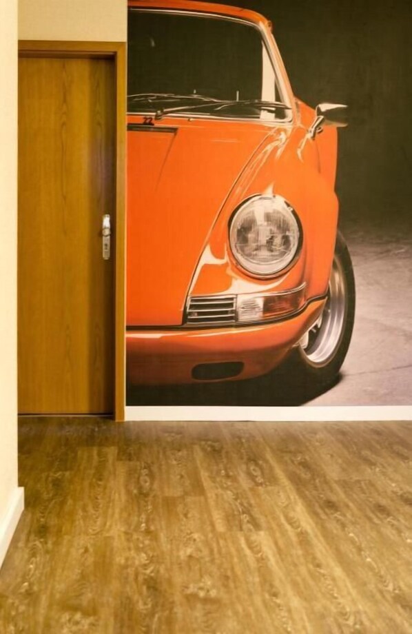 A room with a wooden floor and a door leading to a space with a large poster of an orange vintage car. - Reinhardts Garni Automobilia Hotel (Bietigheim-Bissingen)