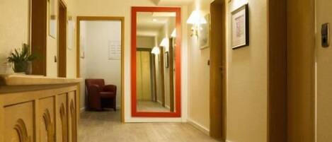 A hotel corridor with wooden flooring, a mirror on the wall, and a red-framed door at the end.