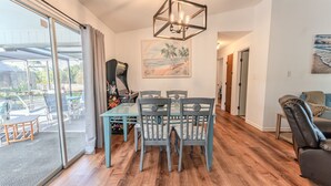 A dining area with a glass table and chairs, a painting of a beach scene, and a view through the glass door to an outdoor patio. - Charming 2BR Home w/Heated Pool & Arcade in Quiet Area – Perfect Location! (Port Charlotte)