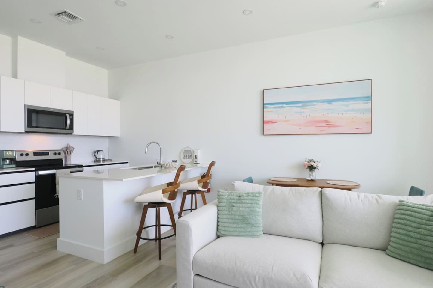 Eine moderne Küche mit Kochinsel, weißen Schränken und einer Mikrowelle. Ein Essbereich mit weißem Sofa und kleinem Tisch ist ebenfalls vorhanden. An der Wand hängt ein Gemälde mit Strandmotiv.