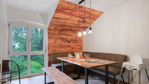 Um interior moderno com uma parede de destaque em madeira, uma mesa de jantar, um banco, um sofá, uma cadeira alta e uma janela grande com vista para o verde.
