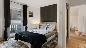Um quarto com cama, janela com cortinas, mesa de cabeceira com abajur e vista para uma varanda.