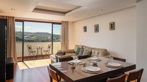 A dining area with a table set for four, a sofa with cushions, and a balcony with a view of the city. - Lobos Horizon – Coziness with Madeiran Soul (Câmara de Lobos)