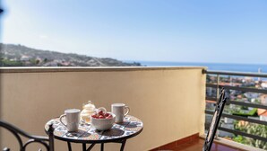 A round table with a patterned tablecloth, two white mugs, a bowl with fruit, and a teapot on a balcony overlooking a coastal town. - Lobos Horizon – Coziness with Madeiran Soul (Câmara de Lobos)